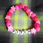 Pink beaded bracelet with letter beads spelling 'Bitch' on a plaid fabric background