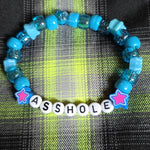 Blue beaded bracelet with letter beads spelling 'ASSHOLE' on a plaid fabric background