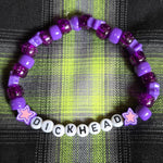 Purple beaded bracelet with 'DICKHEAD' beads on a plaid fabric background