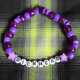 Purple beaded bracelet with 'DICKHEAD' beads on a plaid fabric background